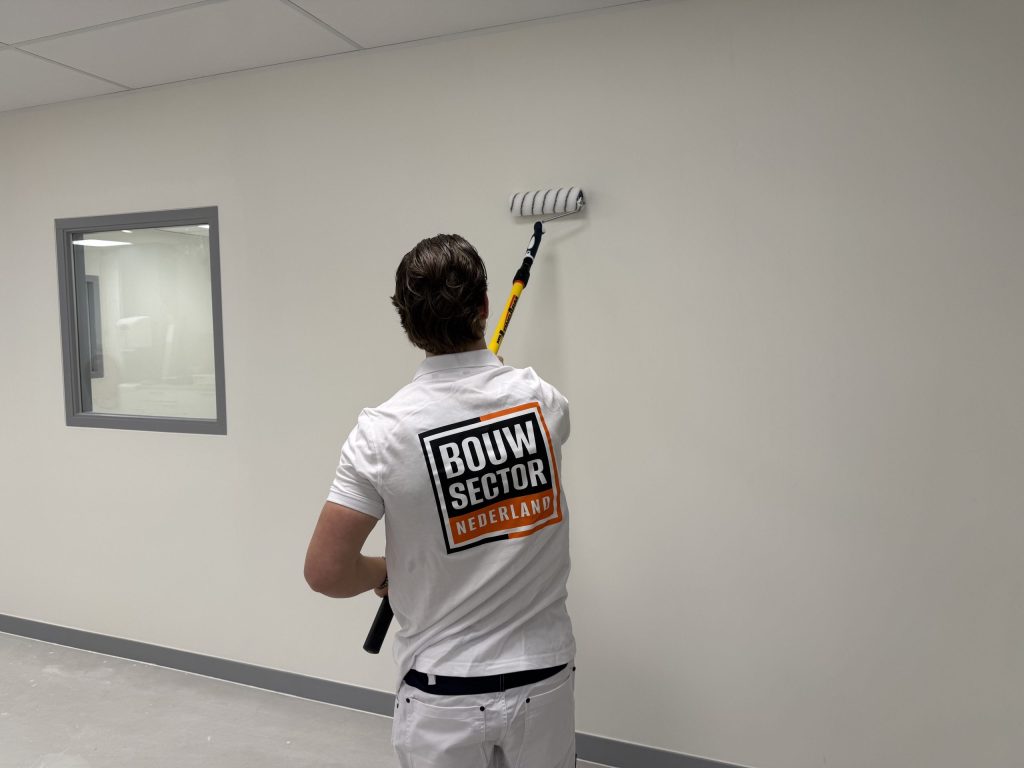 Schilder rolt een witte wand bovenaan naast een raam met grijs kozijn