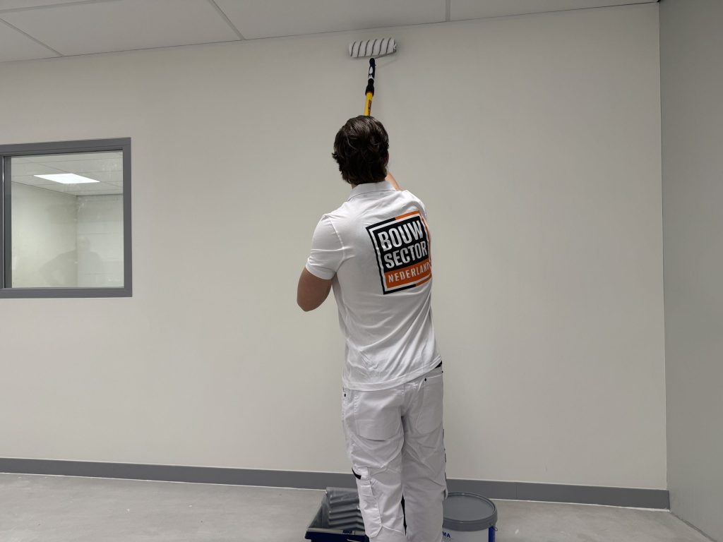 Schilder rolt de bovenrand van een lichte wand tot tegen het plafond, met verfbak en emmer aan zijn voeten