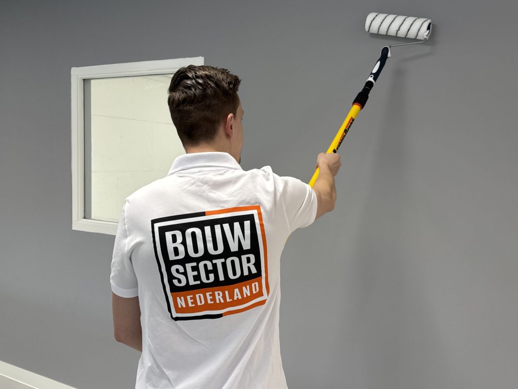 Schilder in een wit poloshirt verft een grijze wand vlak naast een wit gelakt raamkozijn